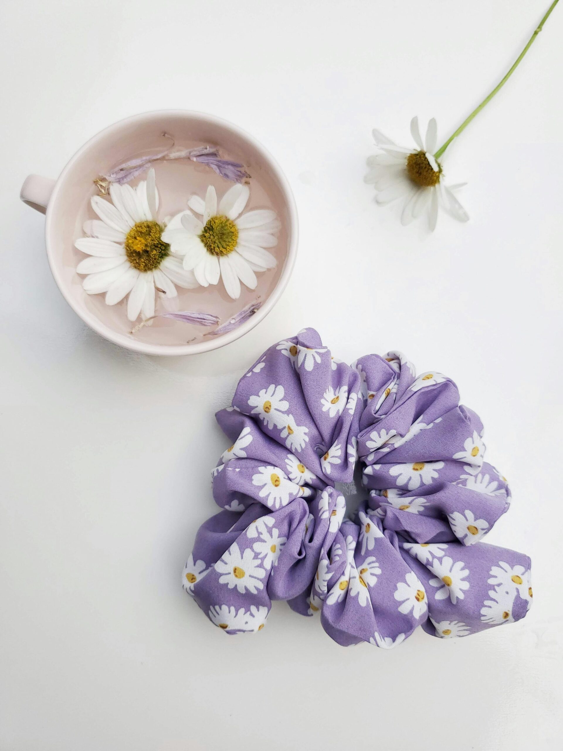 Purple daisy-patterned scrunchie with floating white daisies in pink cup on white background.